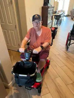 A client drives a mobility scooter through a wide doorway onto a level floor, with a wheelchair parked in the open living area behind him.