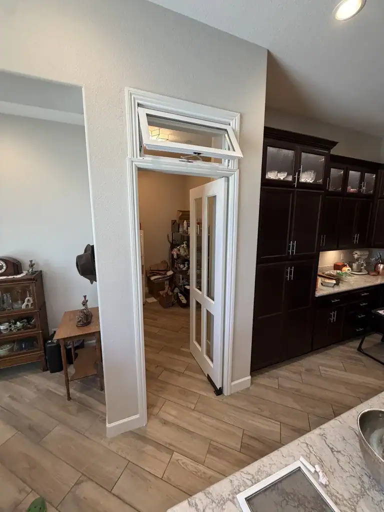 A glass-panel pantry door on double-action hinges pushed open inward, with an operable transom window above it, in a remodeled kitchen.